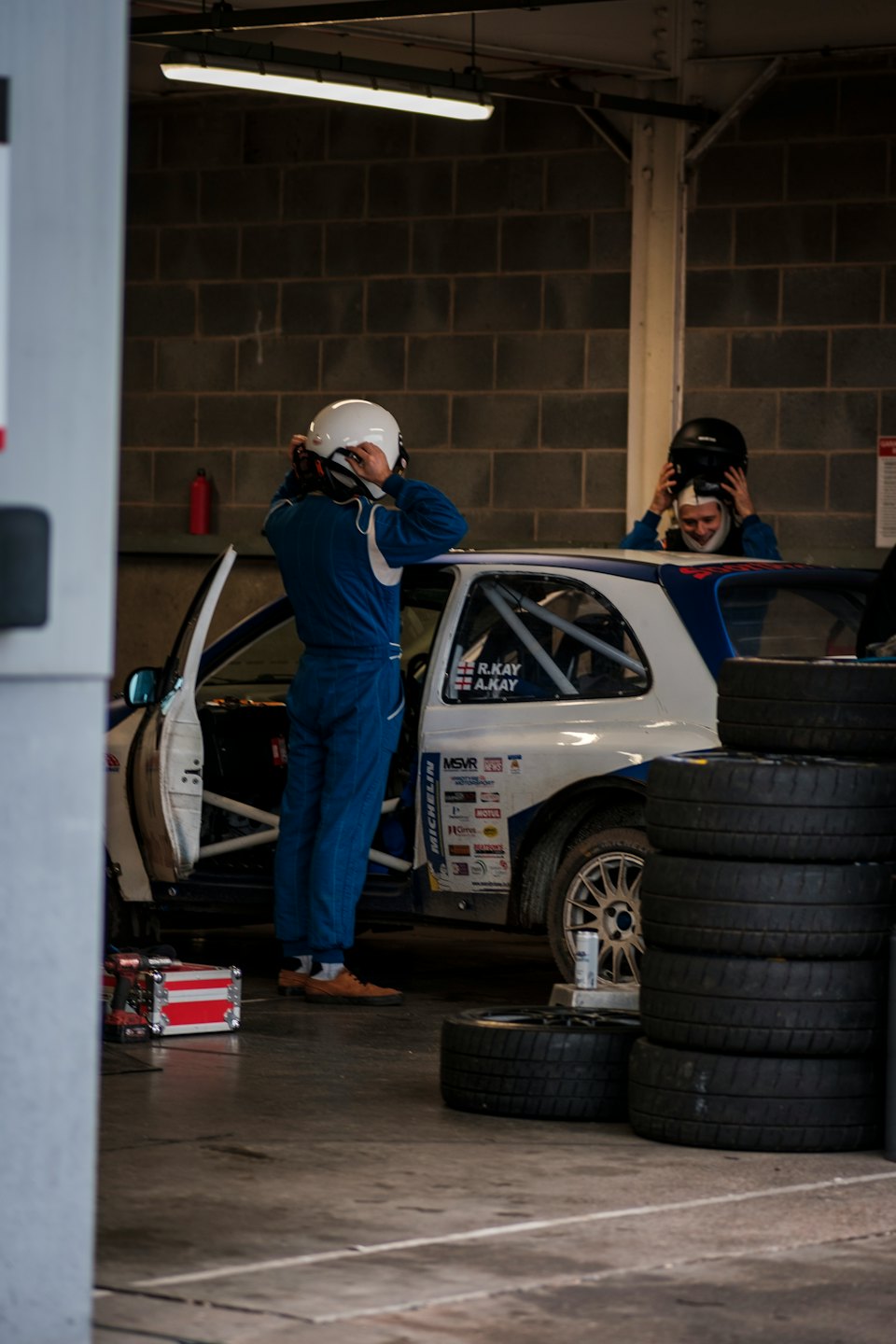 Two racers in blue suits prepare a rally car in a garage.